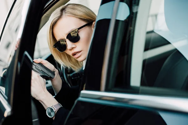 serious blonde woman in sunglasses holding gun in car