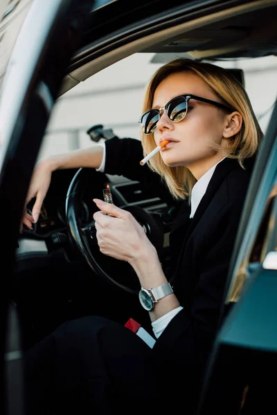 blonde woman in sunglasses holding lighter while smoking cigarette in car