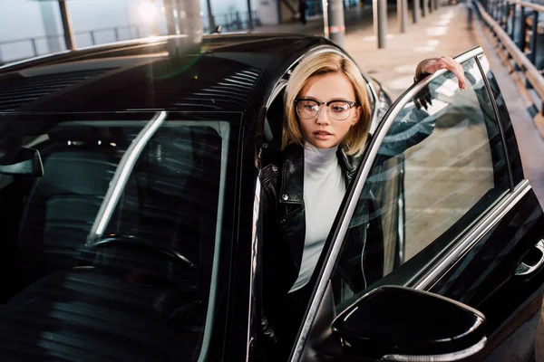 attractive girl in glasses near black automobile in parking