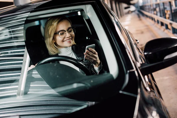 Mujer Atractiva Alegre Gafas Mirando Teléfono Inteligente Mientras Sostiene Volante — Foto de Stock