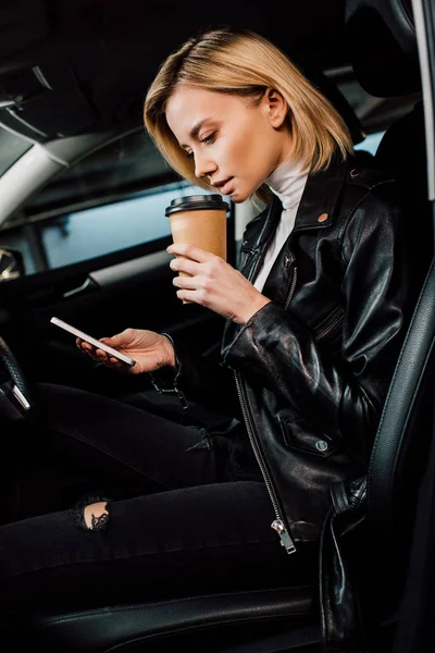 金发女人拿着一次性杯子 在汽车中使用智能手机 — 图库照片
