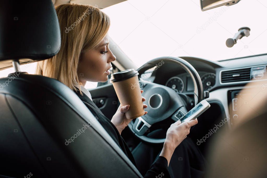 blonde young woman holding paper cup while looking at smartphone in car 