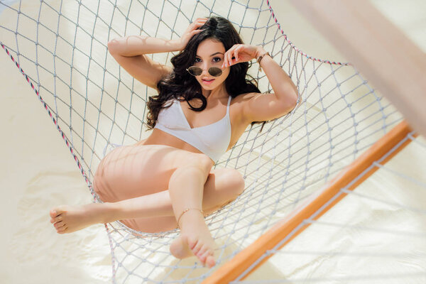 beautiful girl in bikini and sunglasses lying in hammock and looking at camera on beach