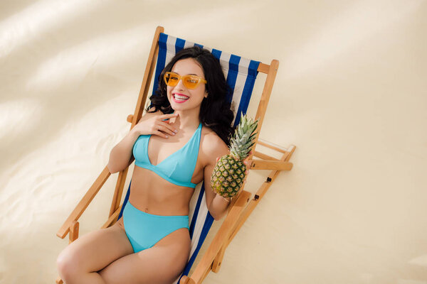 beautiful smiling girl in sunglasses and bikini holding pineapple and relaxing on deck chair on beach
