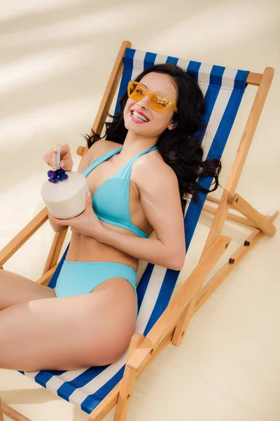 デッキチェアに横たわり ビーチで微笑むココナッツカクテルとビキニとサングラスの美しい女の子 — ストック写真