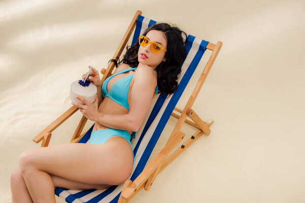beautiful girl in bikini and sunglasses with coconut cocktail lying on deck chair and looking at camera on beach