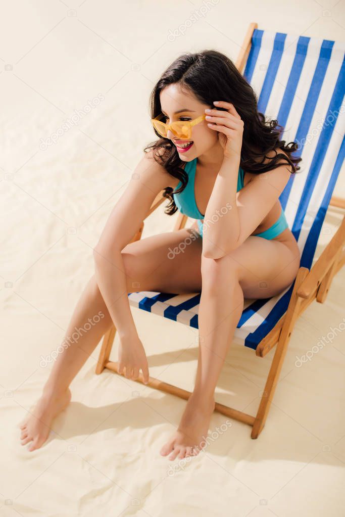 beautiful smiling girl in bikini and sunglasses sitting on deck chair on beach