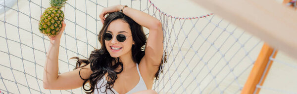 panoramic shot of beautiful smiling girl in sunglasses holding pineapple and lying in hammock on beach 