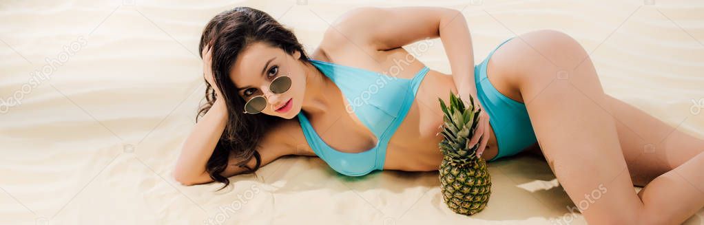 panoramic shot of beautiful girl in bikini and sunglasses with pineapple Looking At Camera while lying on beach