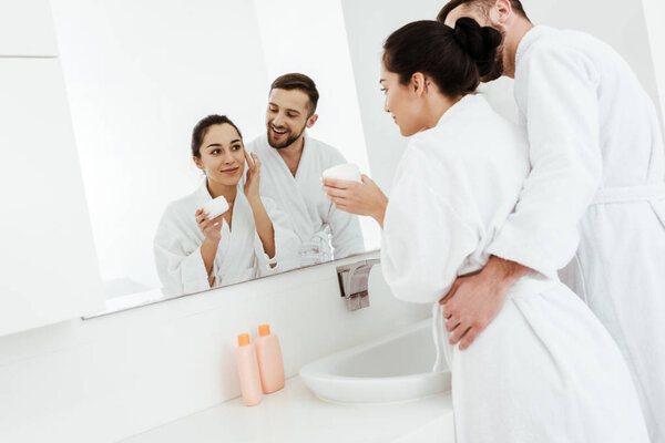 cheerful man looking at beautiful girlfriend applying face cream in bathroom 