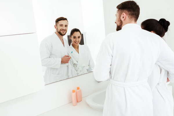 selective focus of bearded man holding toothbrush and looking at mirror with happy brunette woman 