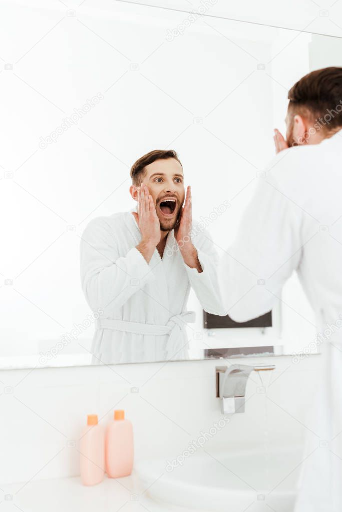 selective focus of handsome bearded man looking at mirror and screaming in bathroom 