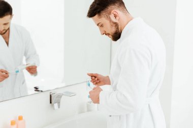 banyoda diş macunu ve diş fırçası tutan yakışıklı sakallı adam seçici odak 