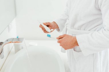 cropped view of man holding toothbrush and toothpaste near sink in bathroom  clipart