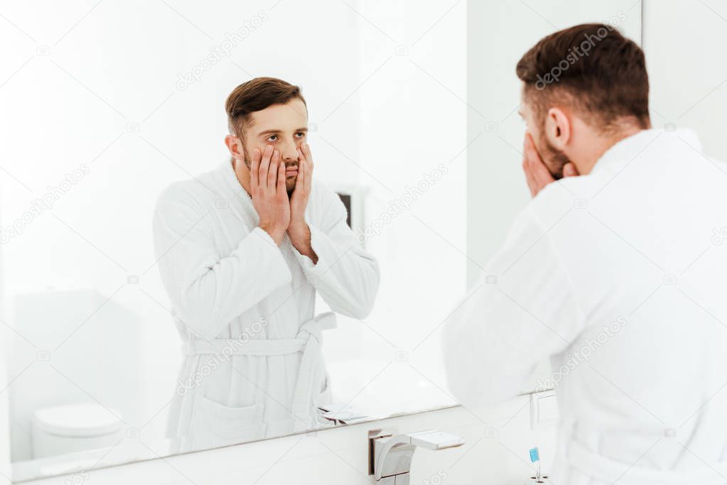 selective focus of upset bearded man looking at mirror while touching face in bathroom 