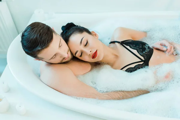 overhead view of handsome man lying in bathtub with sexy brunette woman in lace bra