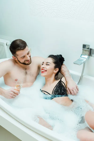 シャンパングラスを持ち 浴槽で幸せな女性を見ている陽気な男 — ストック写真