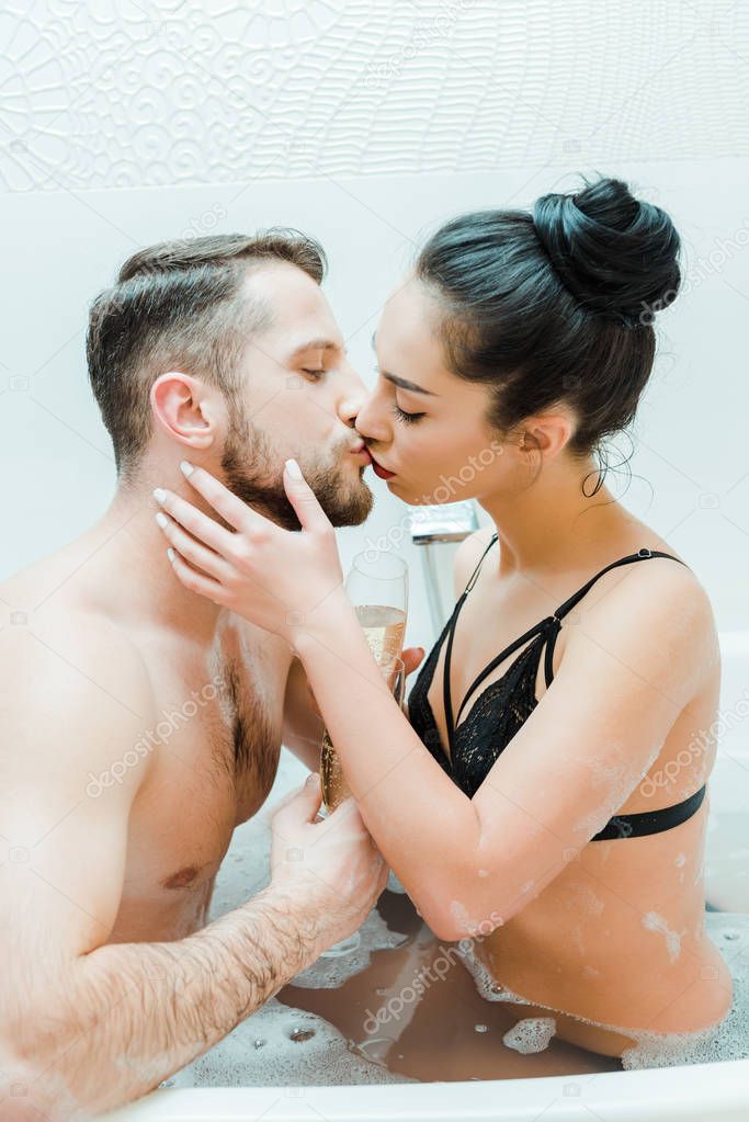 handsome man kissing with sexy young woman holding champagne glasses in bathtub 
