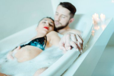 banyo köpüğü ile küvette yatarken yakışıklı adam ve çekici esmer kadın el ele tutuşan seçici odak 