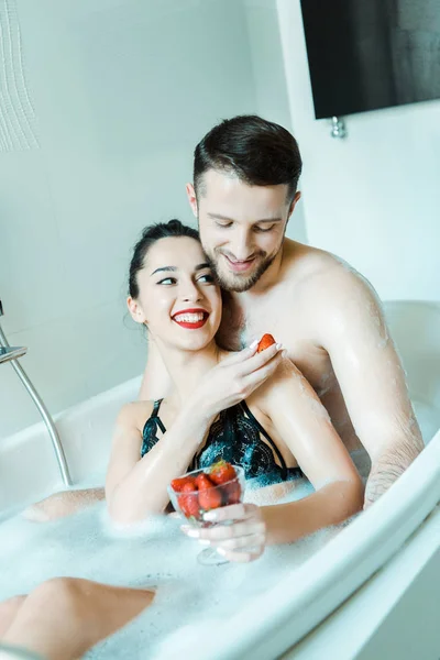 快乐的年轻女子拿着美味的草莓附近的欢快的男朋友在浴缸 — 图库照片
