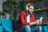 pohled s nízkým výhledem na šťastného vousatého muže naslouchající hudbu ve sluchátkách a používání smartphone v obývacím pokoji 