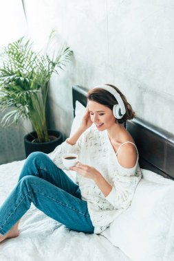 yatakta kahve bir fincan tutarken kulaklık müzik dinleyen neşeli kadın havai görünümü 