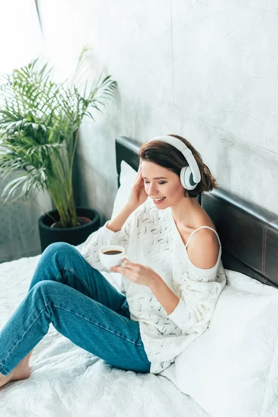 欢快的女人在床上喝咖啡时在耳机里听音乐的头顶视图 — 图库照片