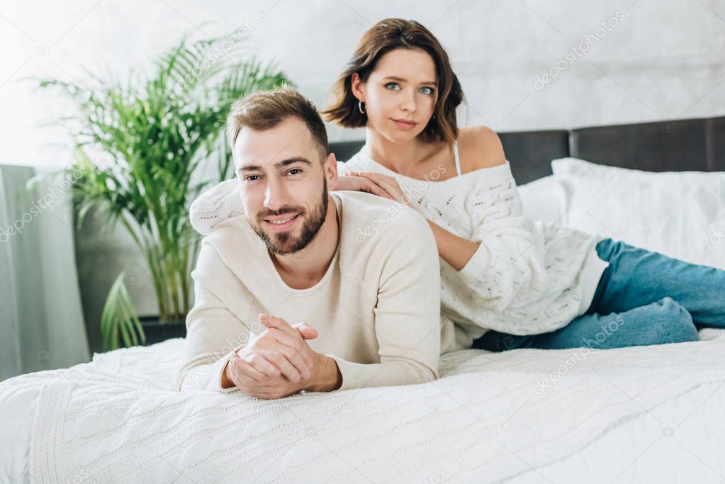 happy bearded man with clenched hands looking at camera with attractive woman while lying on bed 