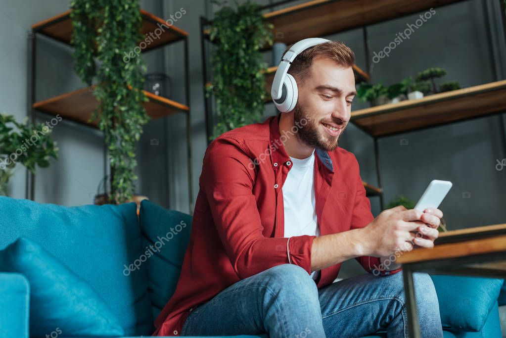 Low angle view of happy bearded man listening music in headphones and using smartphone in living room