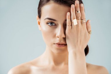 young naked woman with shiny makeup and golden rings hiding face behind hand isolated on grey clipart