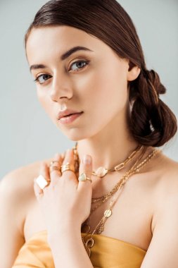 young attractive woman with shiny makeup in golden necklaces and rings looking at camera isolated on grey clipart