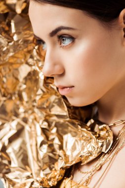 young woman with shiny makeup and golden foil in necklaces looking away clipart