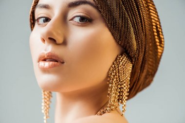 young beautiful woman with shiny make up, turban and golden earrings looking at camera isolated on grey clipart