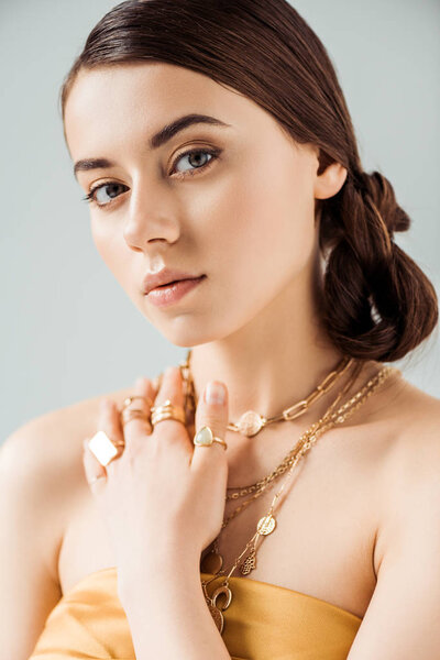 young attractive woman with shiny makeup in golden necklaces and rings looking at camera isolated on grey