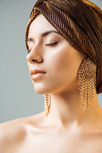 young naked woman with closed eyes, shiny makeup, golden rings in turban isolated on grey