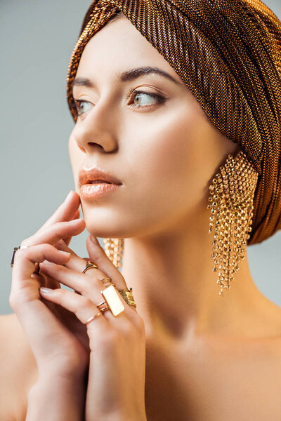 young naked woman with shiny makeup, golden rings and earrings in turban looking away isolated on grey