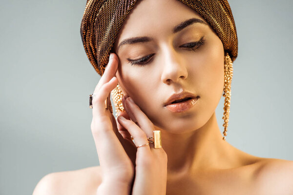 young nude woman with hands near face, shiny makeup, golden rings and earrings in turban isolated on grey