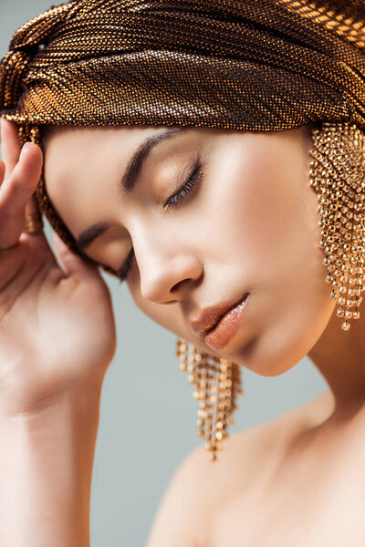 young woman with closed eyes, shiny makeup and golden earrings in turban isolated on grey