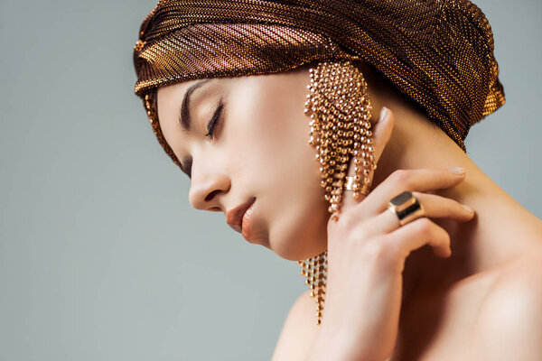 young nude woman with closed eyes, shiny makeup, golden rings and earrings in turban touching neck isolated on grey