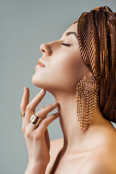 profile of young nude woman with closed eyes, shiny makeup, golden rings and earrings in turban touching neck isolated on grey