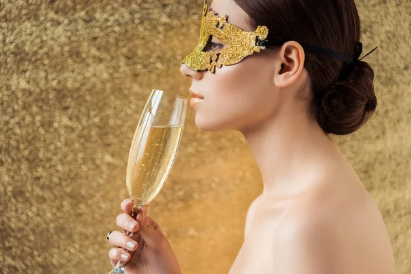 光沢のあるメイクとシャンパンのガラスで金色のマスクを着けた若い裸の女性のプロフィールは テクスチャードの黄金の背景にカメラを見て — ストック写真