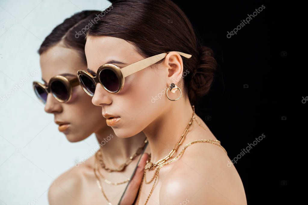 young naked woman in sunglasses, golden accessories holding mirror isolated on black