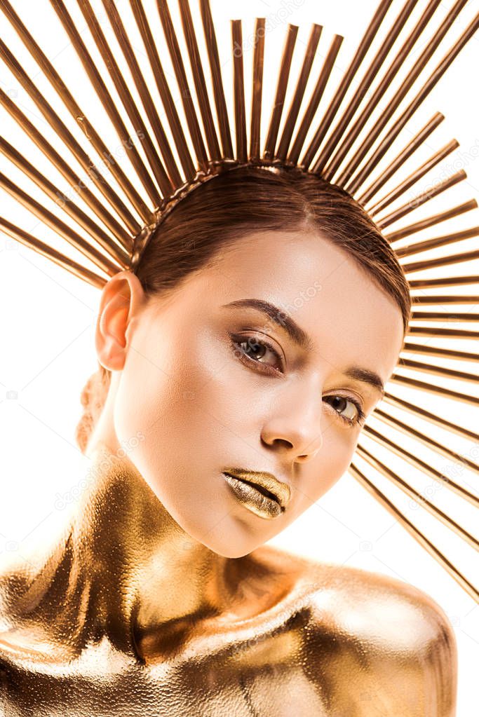 naked young woman painted in golden with accessory on head looking at camera isolated on white