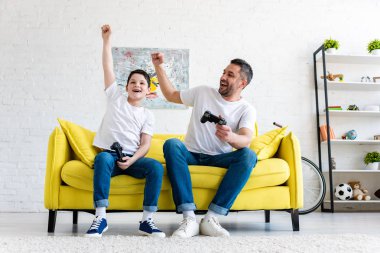 father and son cheering while playing Video Game on couch at home clipart