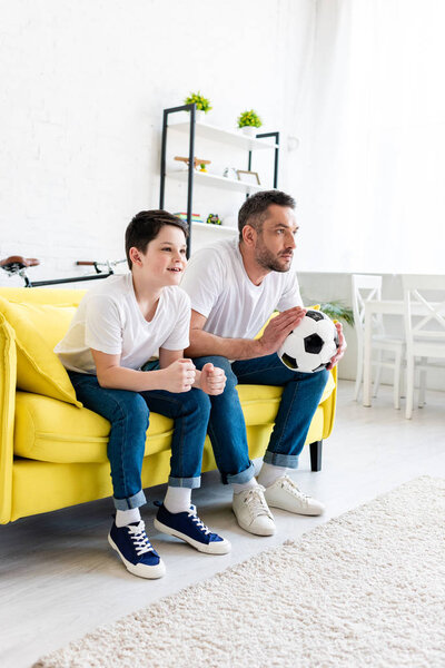 отец и сын смотрят спортивный матч дома на диване
