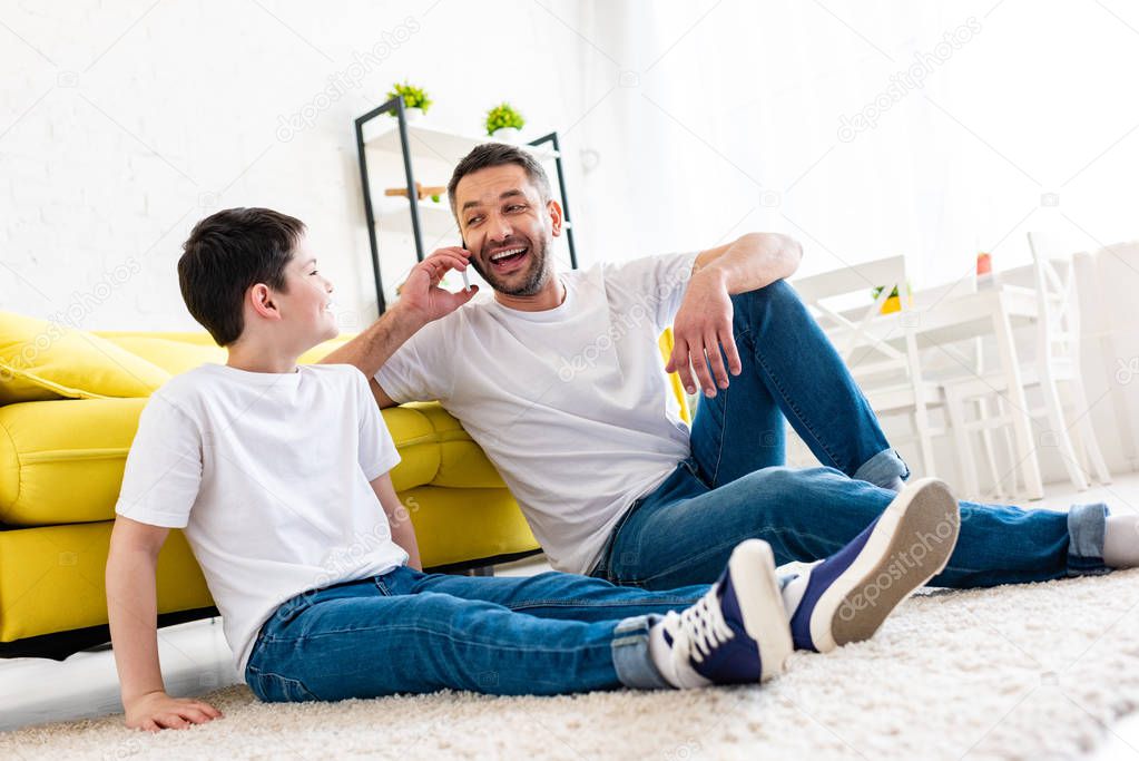 son sitting on carpet near happy father talking on smartphone in Living Room