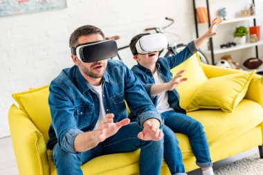father and son in vr headsets experiencing Virtual reality on couch at home clipart