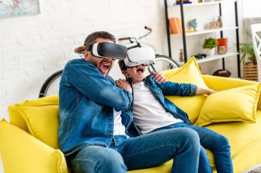 father and son in vr headsets yelling while experiencing Virtual reality on couch at home clipart