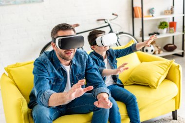 father and son in vr headsets experiencing Virtual reality on couch at home clipart