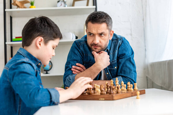 handsome father and son in denim playing chess while sitting at table at home